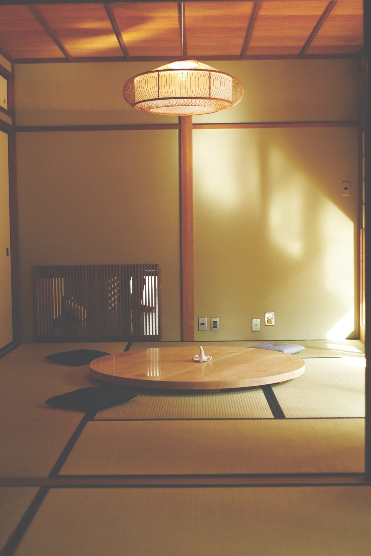 Minimalist Japanese room with tatami mats, round wooden table, woven pendant lamp, and traditional wooden screen divider.
