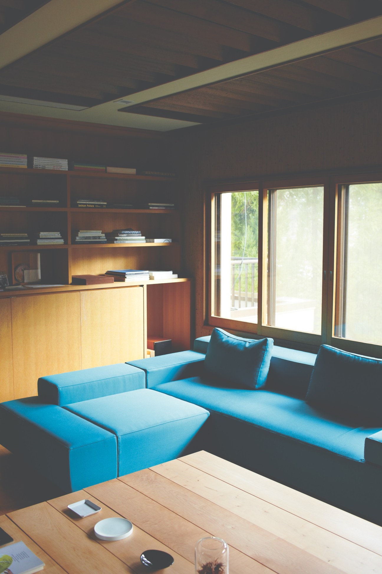 Modern office space with turquoise sofa, wood desk, built-in shelving, and large windows overlooking gardens.