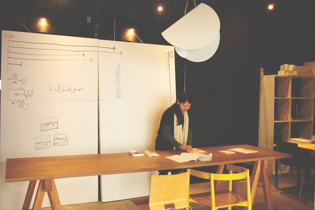 A staff member works at a wooden table in a basement workshop with large white design boards and pendant lighting.