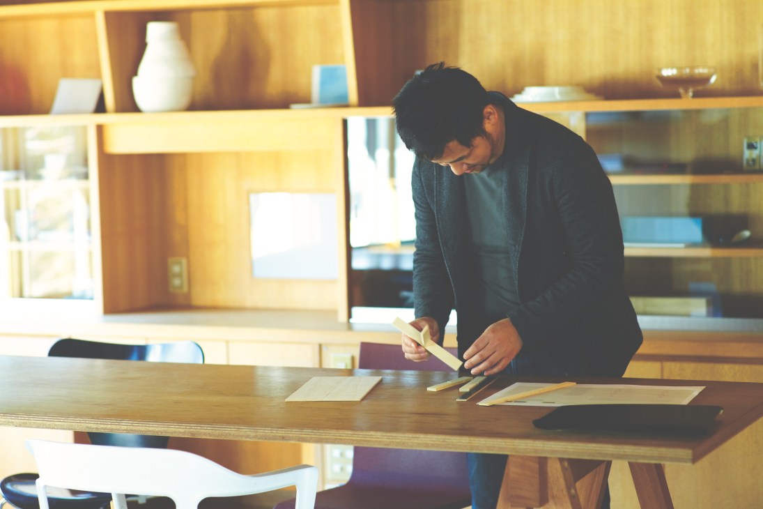 Teruhiro Yanagihara works at a wooden desk in his minimalist office with warm wood paneling and shelving.