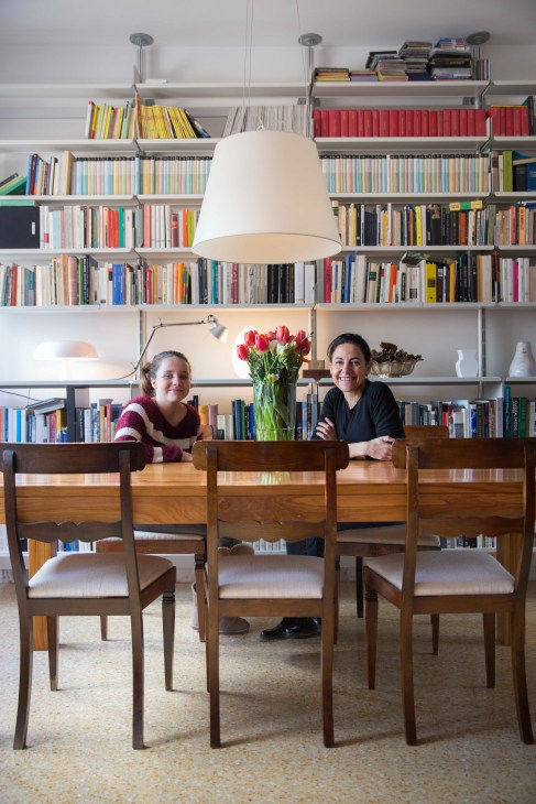 Simona Salvo and her daughter Elena sit at a wooden dining table in a modern living room with floor-to-ceiling bookshelves.