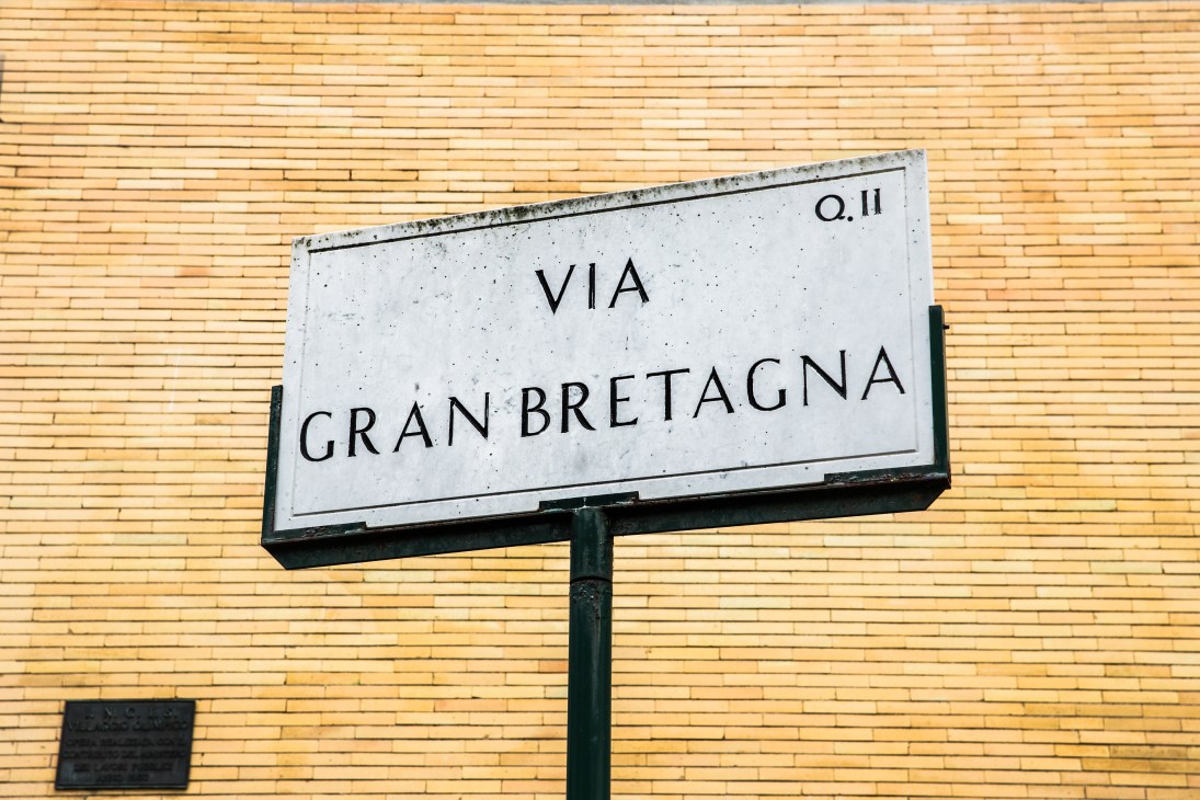 A weathered marble street sign reading 'VIA GRAN BRETAGNA' mounted on a green pole against a yellow brick wall.