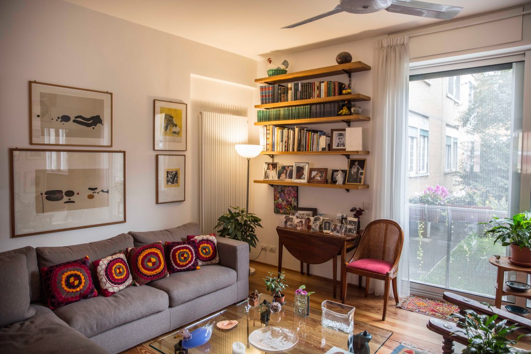 Modern living room with gray sofa, wooden shelving, framed artwork, and views of Rome's Olympic Village courtyard through floor-to-ceiling windows.