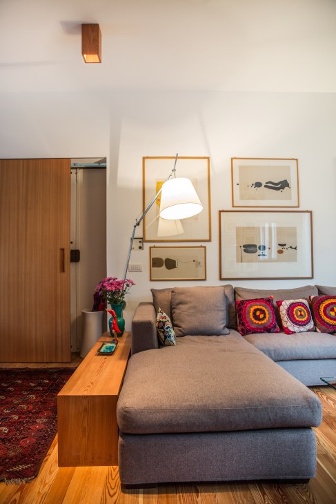 Modern living room with grey sectional sofa, colorful patterned cushions, wooden wardrobe, arc floor lamp, and framed artwork on white walls.