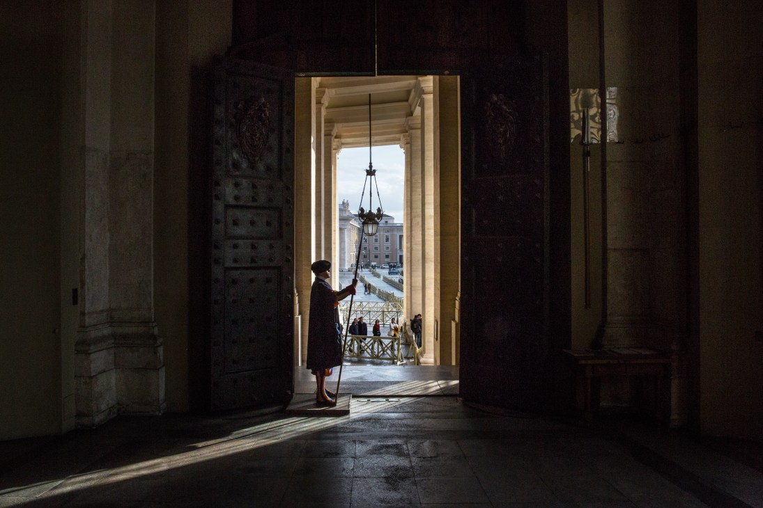 A figure in dark clothing stands in a Vatican loggia doorway looking out toward St Peter's Square.
