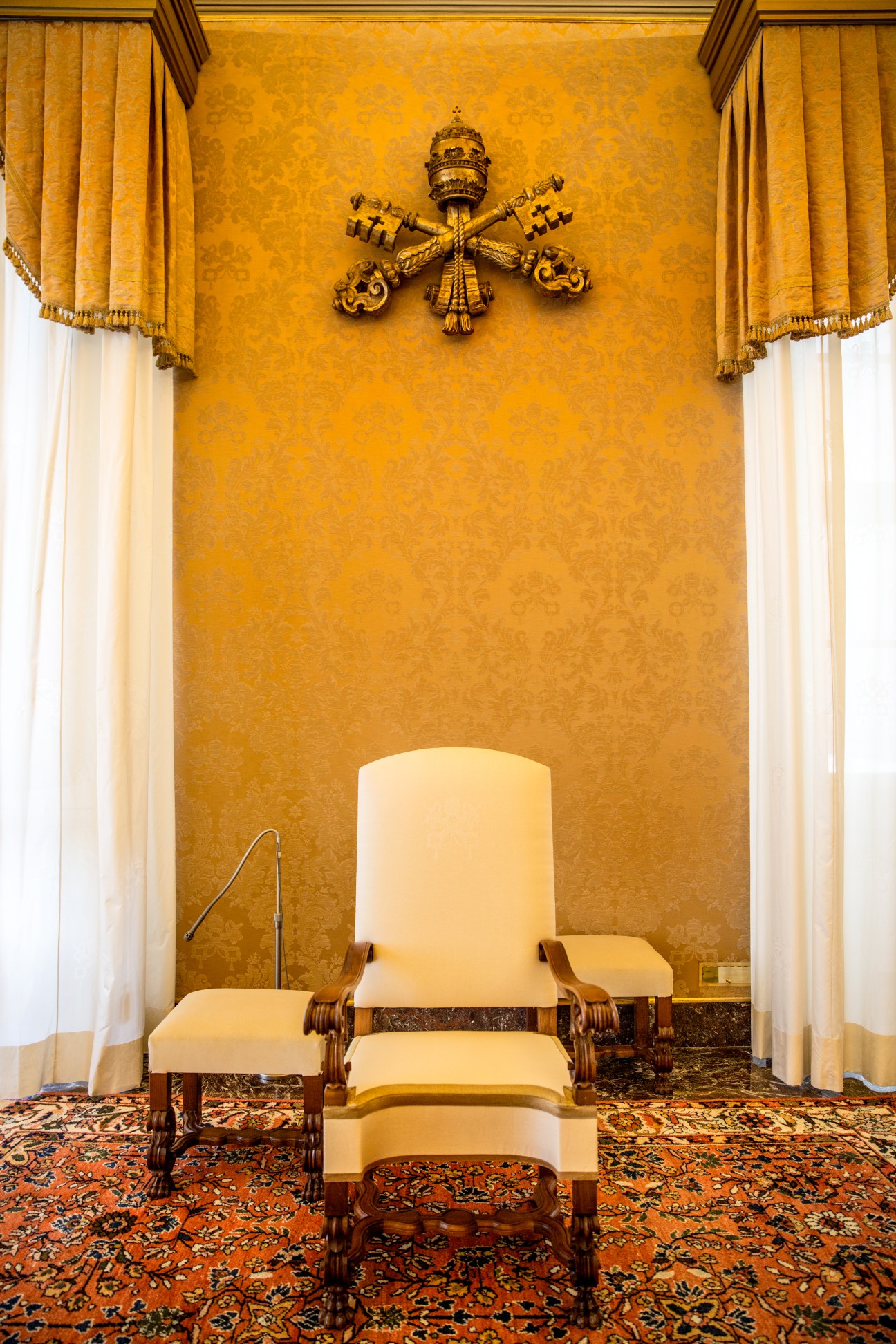 Papal chair with cream upholstery and wooden arms in gold-damask room with papal coat of arms.