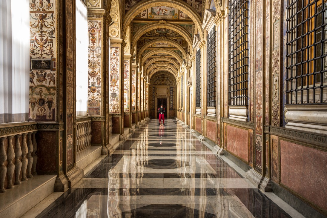 A Swiss guard in red uniform walks through an ornate Vatican loggia with gilded Renaissance frescoes and marble floors.