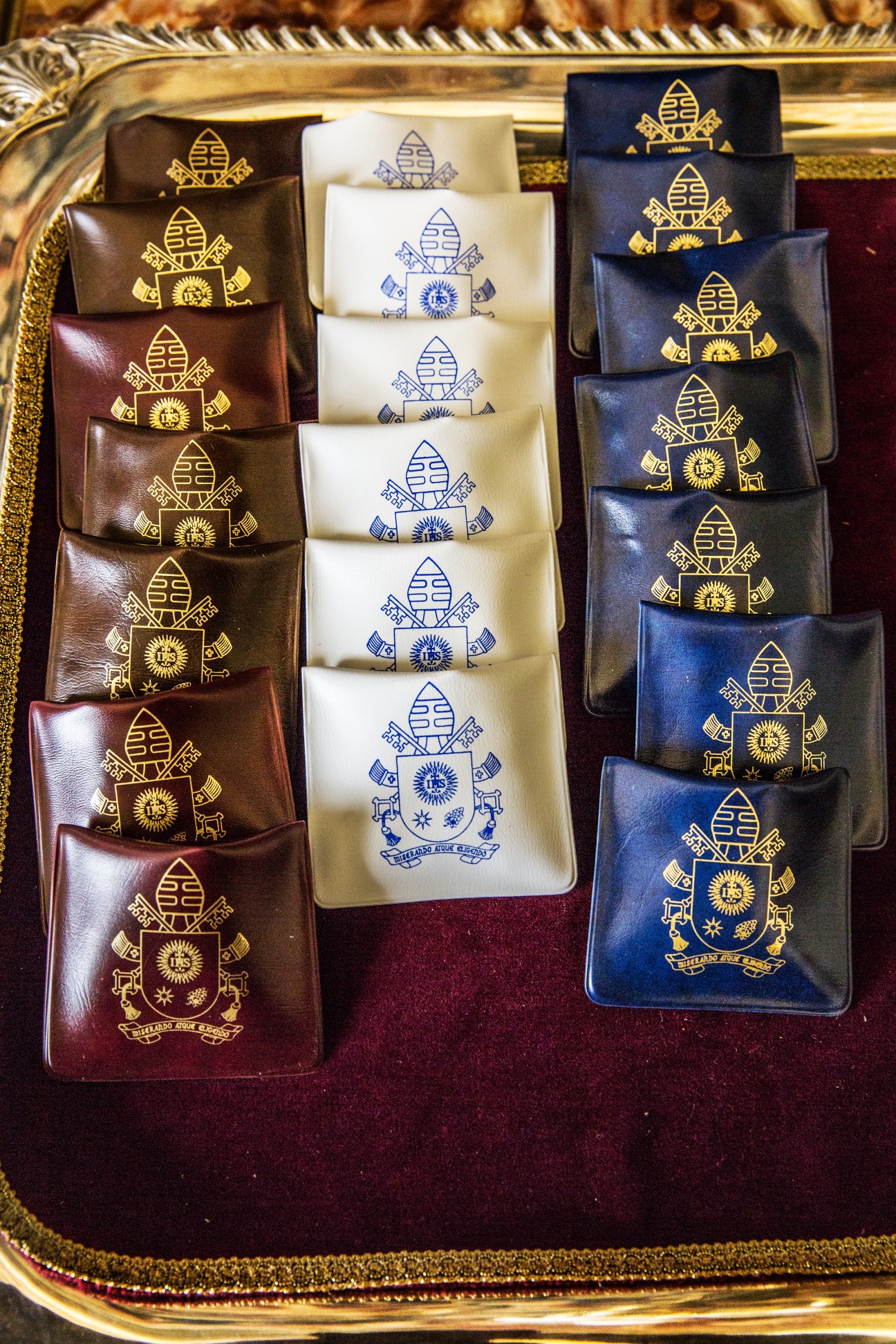 Papal coat of arms embroidered on leather wallets in brown, white and navy blue.