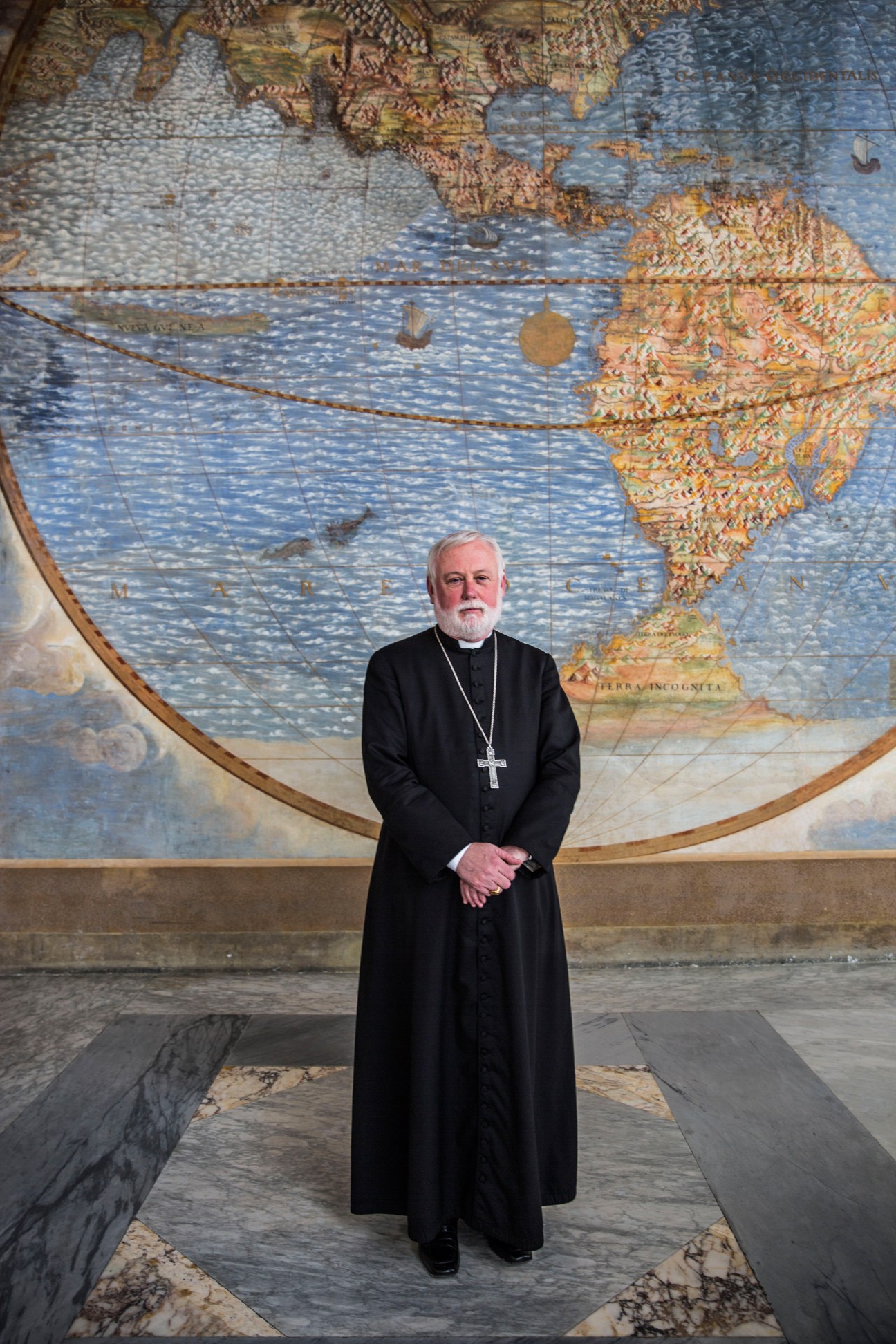Archbishop Paul Gallagher standing in black cassock before a 16th-century world map fresco in the Vatican's Apostolic Palace.