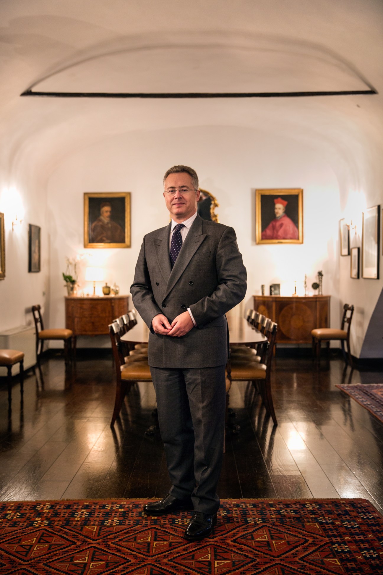 British ambassador Nigel Baker stands in a formal diplomatic residence with classical portraits and period furnishings.