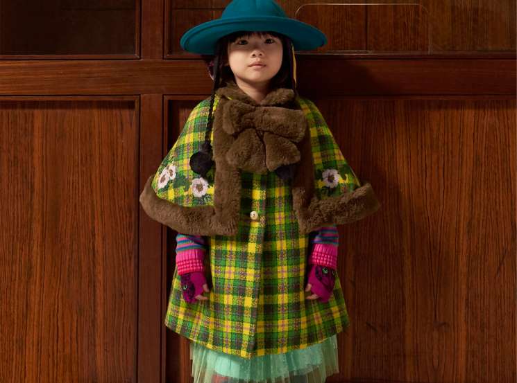 Child wearing colorful Gucci childrenswear including plaid coat, blue hat, and floral boots against wooden paneling
