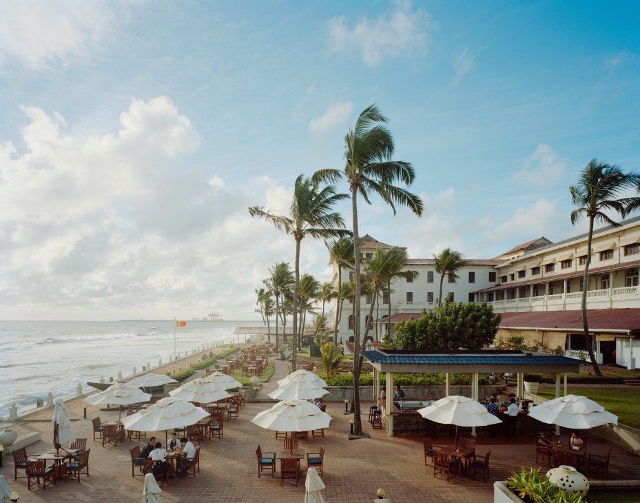 The terrace of the Galle Face Hotel with white umbrellas, wooden chairs, palm trees and the Indian Ocean.