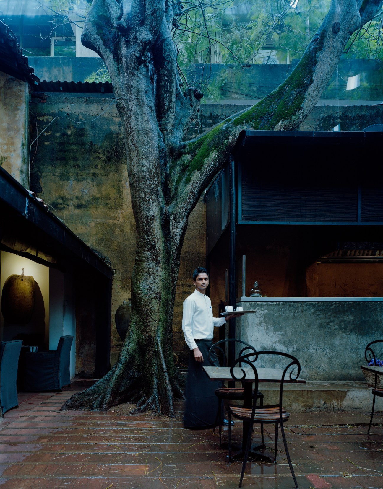 Waiter holding a tray stands in a courtyard with a large ancient tree and weathered colonial architecture.