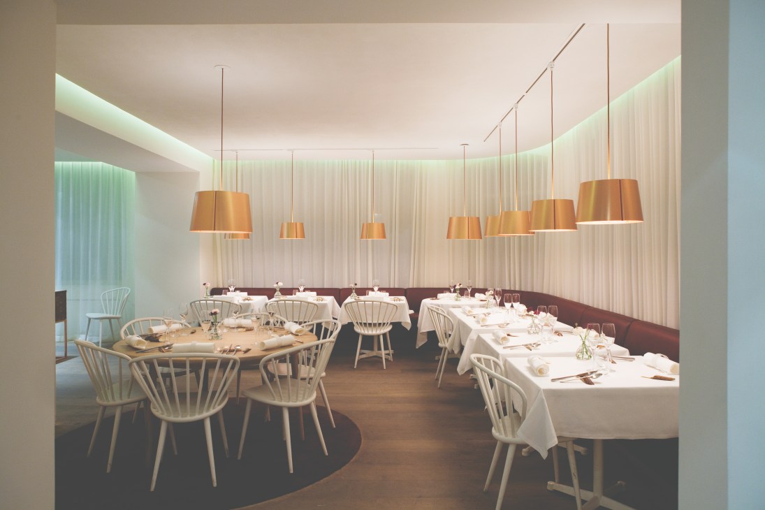 Modern restaurant dining room with white pendant lights, brown leather banquette seating, and Windsor chairs at round tables set for service.