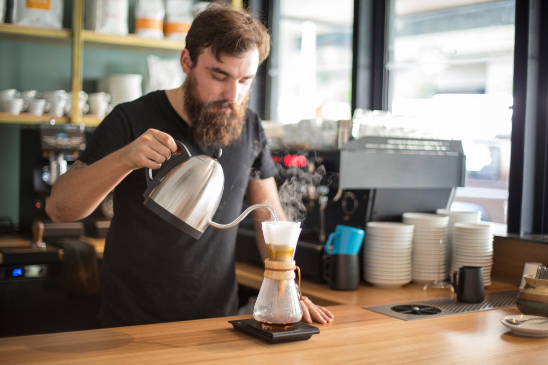 A bearded barista pours hot water from a gooseneck kettle into a pour-over coffee filter at Gauge, Brisbane.