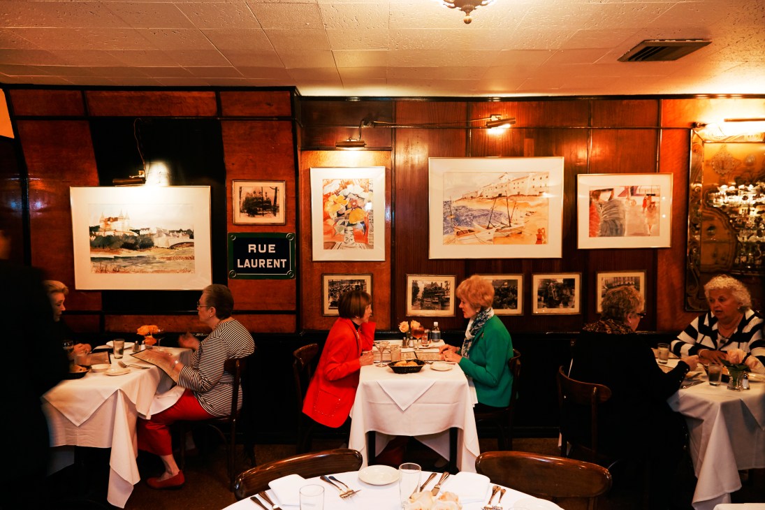 Le Veau d'Or dining room with wood-paneled walls, framed artwork, white-linen tables and seated diners.