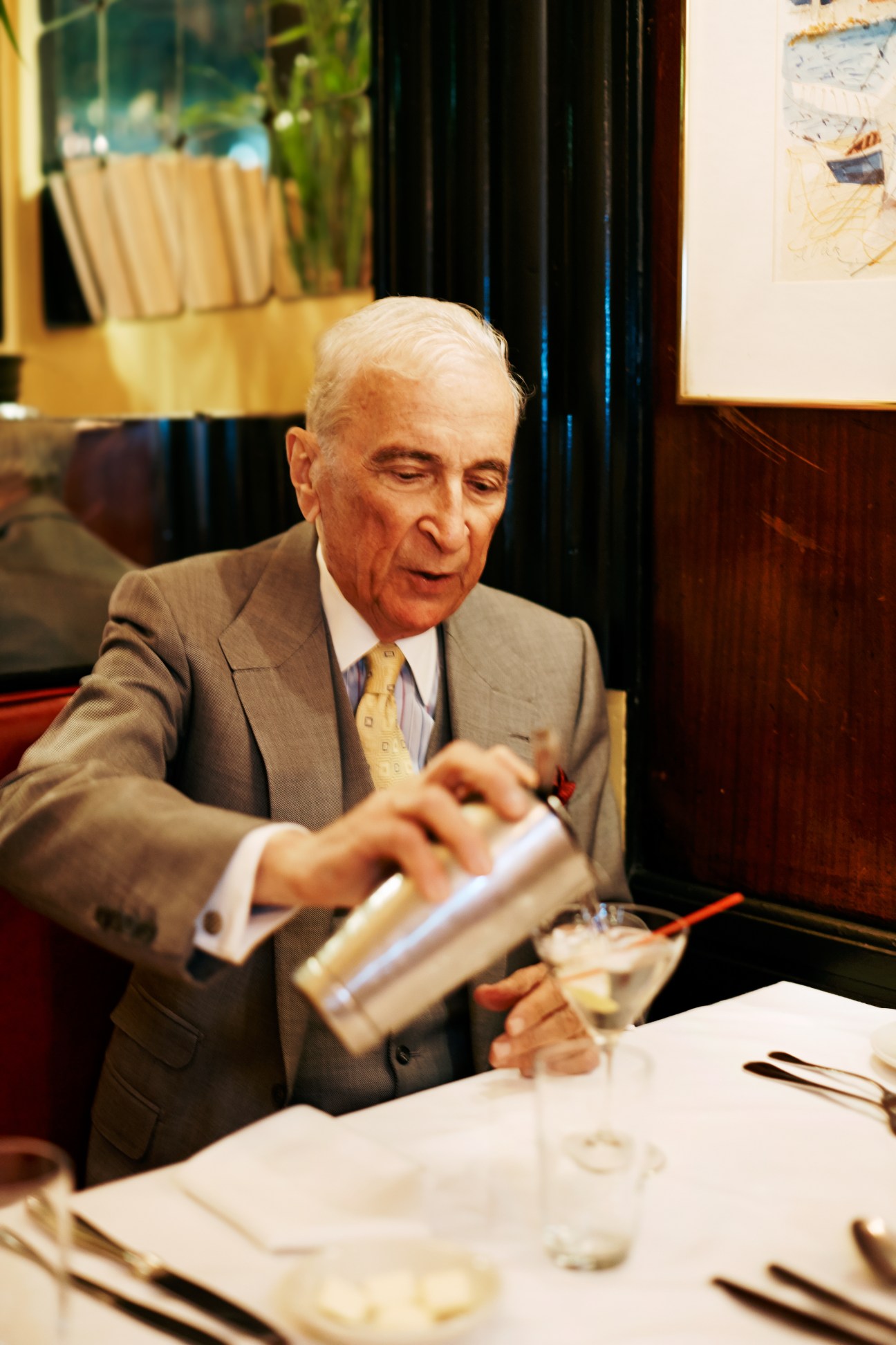 Gay Talese in a grey suit pouring gin into a martini glass at his table at Le Veau d'Or restaurant.