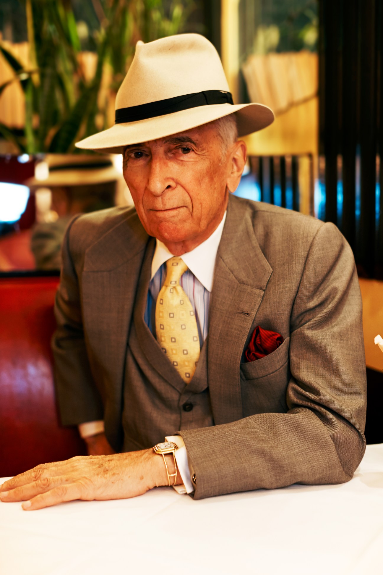 Elegantly dressed older man in panama hat and beige suit seated at restaurant table.