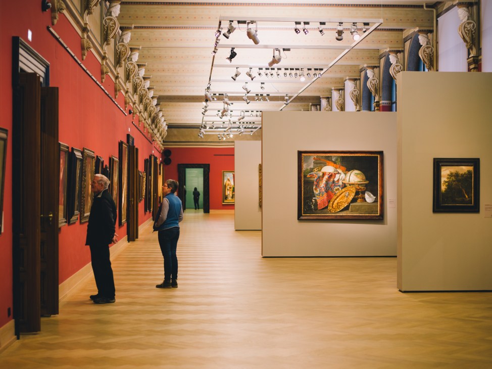 Visitors view old master paintings displayed on red and cream walls in a gallery with ornate ceiling and track lighting.