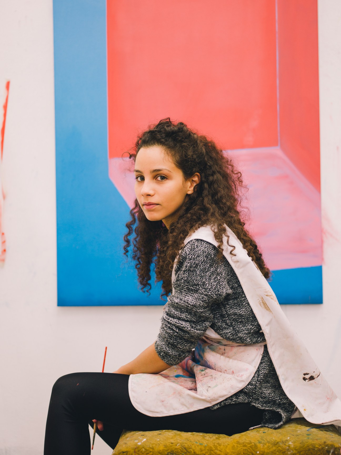 Student sitting on a bench taking a break, wearing paint-stained sleeves against a colorful abstract artwork.