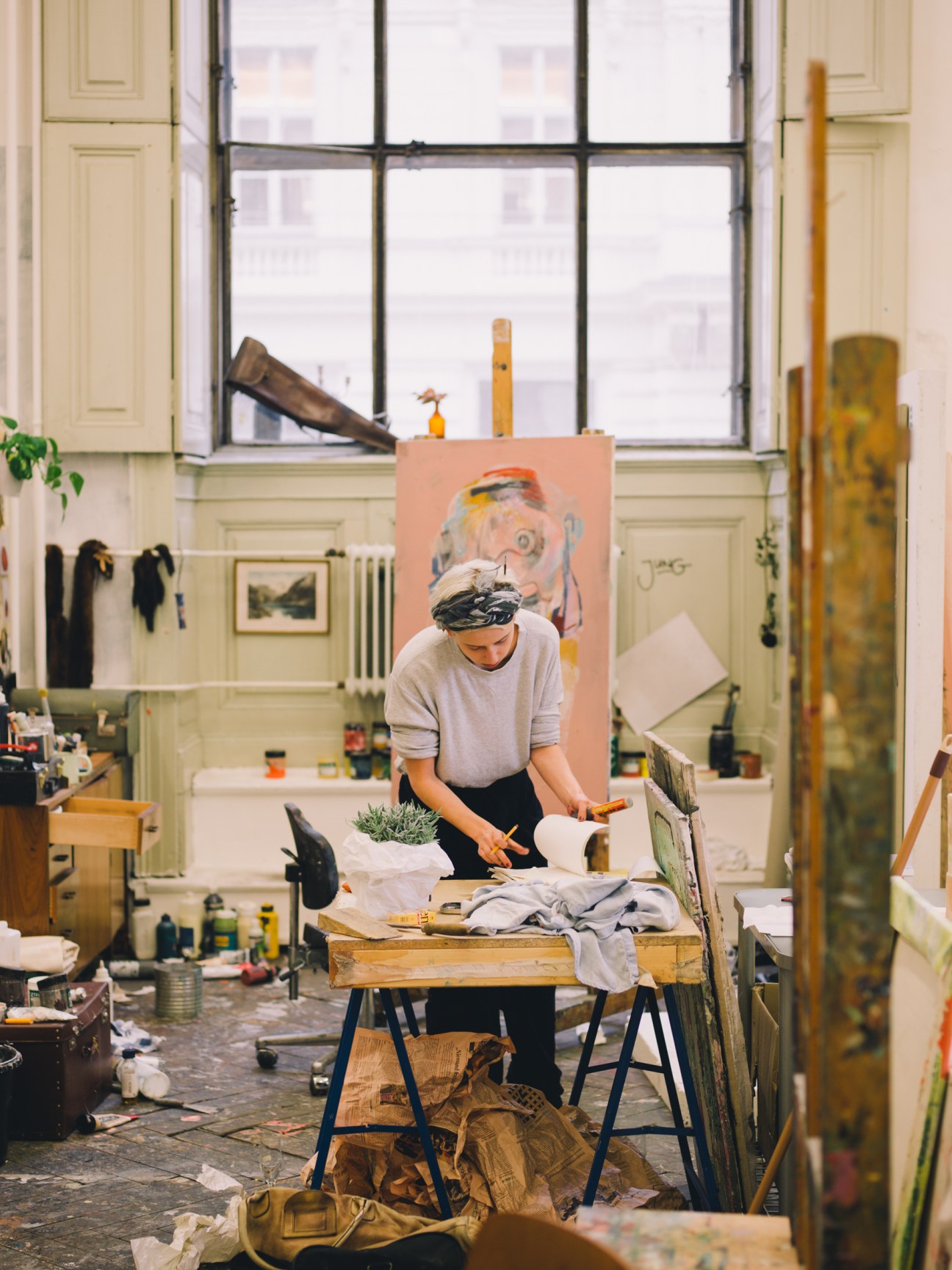 Student working at a table in a bright studio with paint supplies and a canvas on easel behind her.