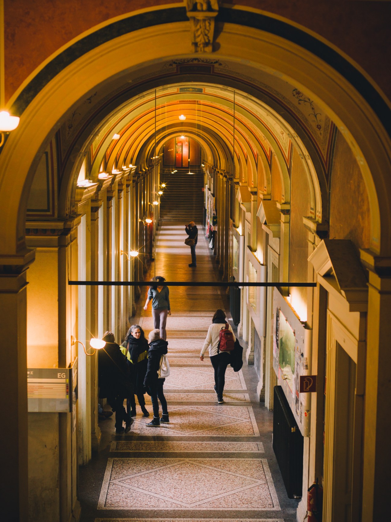 A grand arched hallway with golden walls and vaulted ceiling, lined with tall wooden doors and lit by wall sconces, with students walking through.