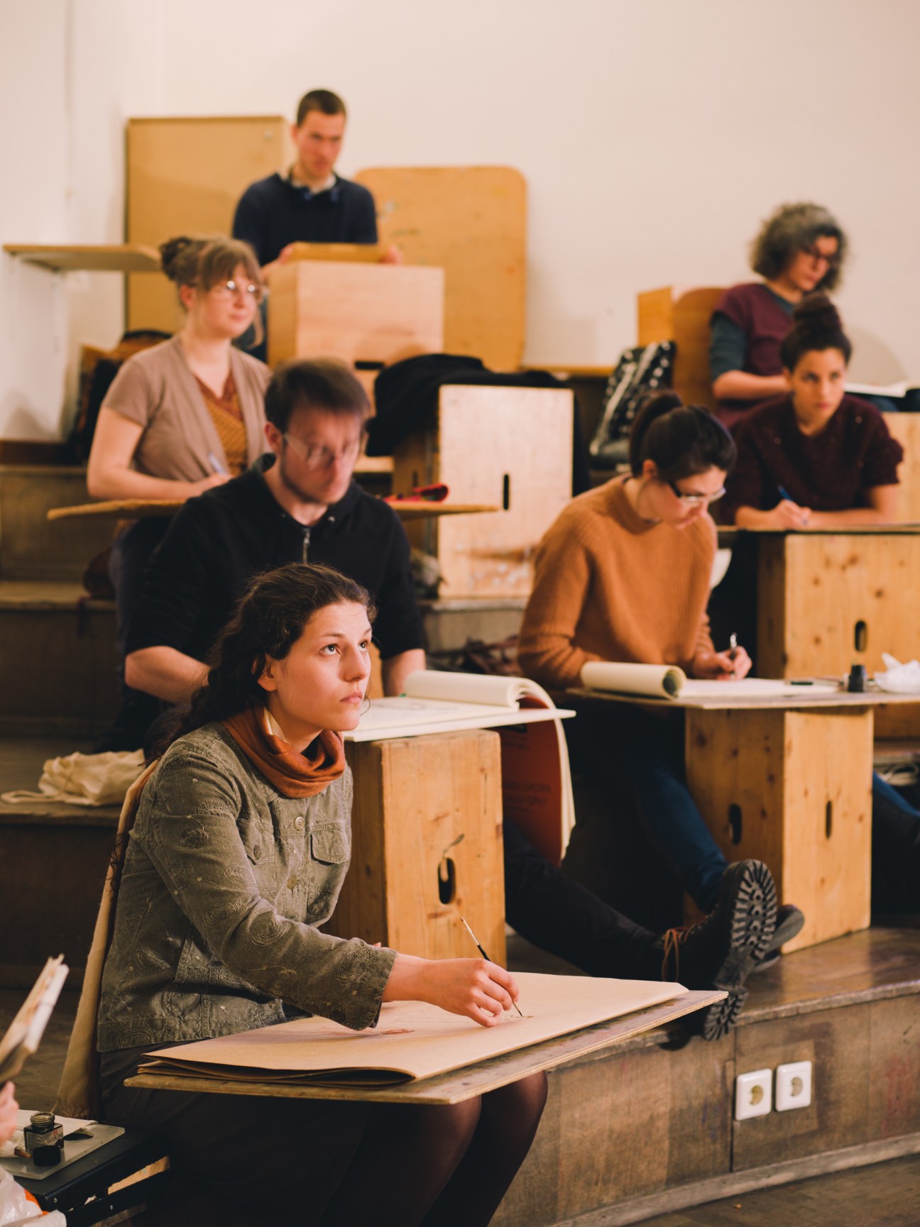 Multiple students sitting at wooden easels in a life-drawing class, focused on their work.