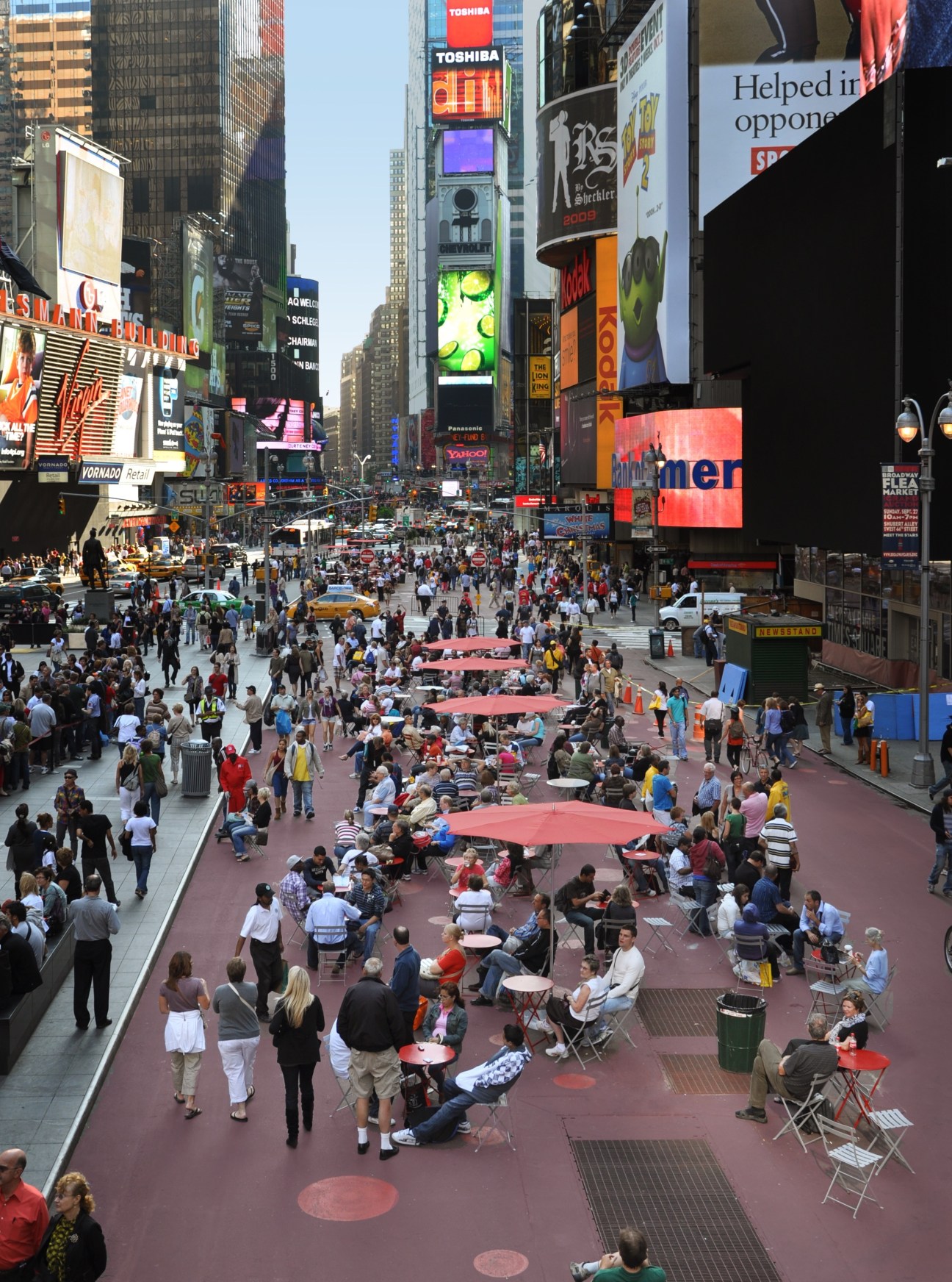Times Square in New York with pedestrians seated at tables and standing in a pedestrianized plaza with red umbrellas and seating areas.