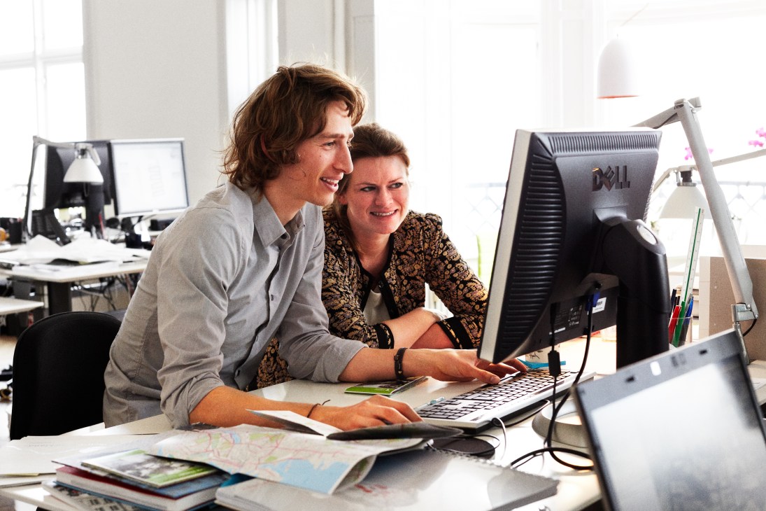 Two colleagues working together at a desk with a computer and city maps in a bright office.