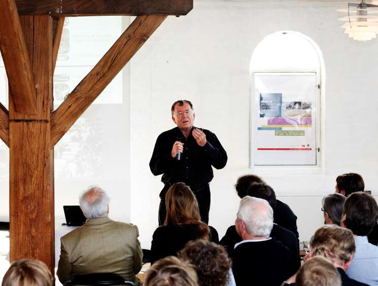 Jan Gehl lecturing at his book launch, speaking to an audience in a modern space with wooden beams and framed architectural posters on the walls.