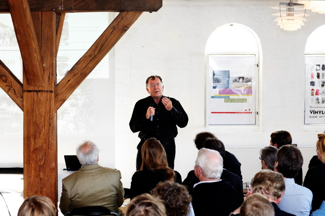 Jan Gehl lecturing at his book launch, speaking to an audience in a modern space with wooden beams and framed architectural posters on the walls.