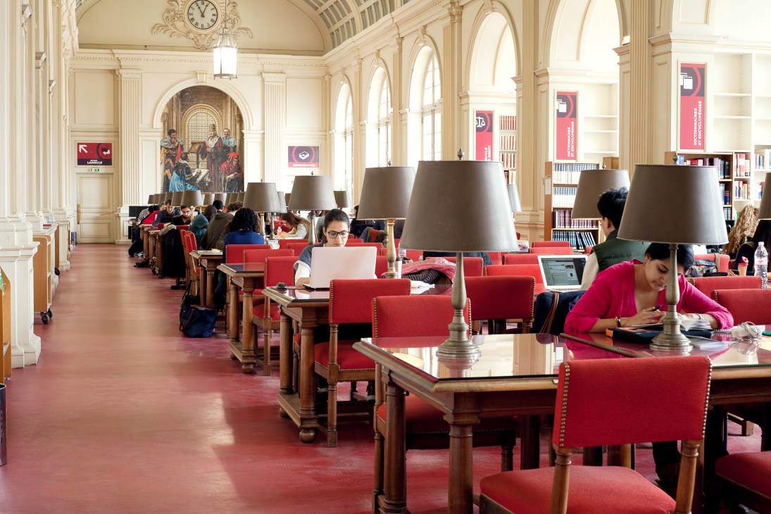 Students studying at desks with red chairs in an ornate library with classical columns and arched windows.