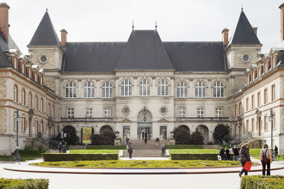 Grand historic building with mansard roof and classical architecture, surrounded by manicured gardens and courtyards with students and visitors.