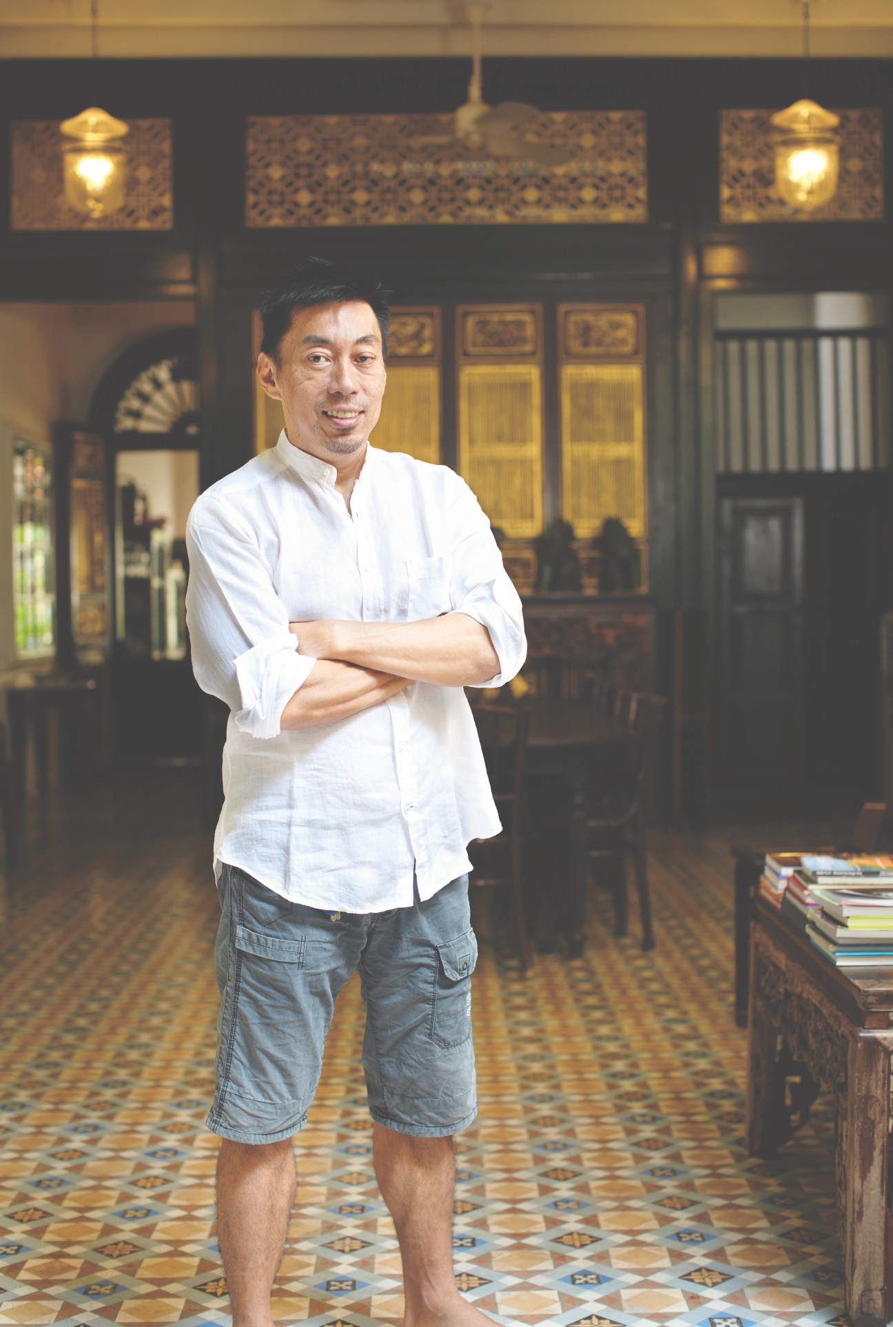 Christopher Ong, a hotelier and entrepreneur, standing with arms crossed in a traditional colonial-style interior with ornate wooden doors and decorative lanterns.