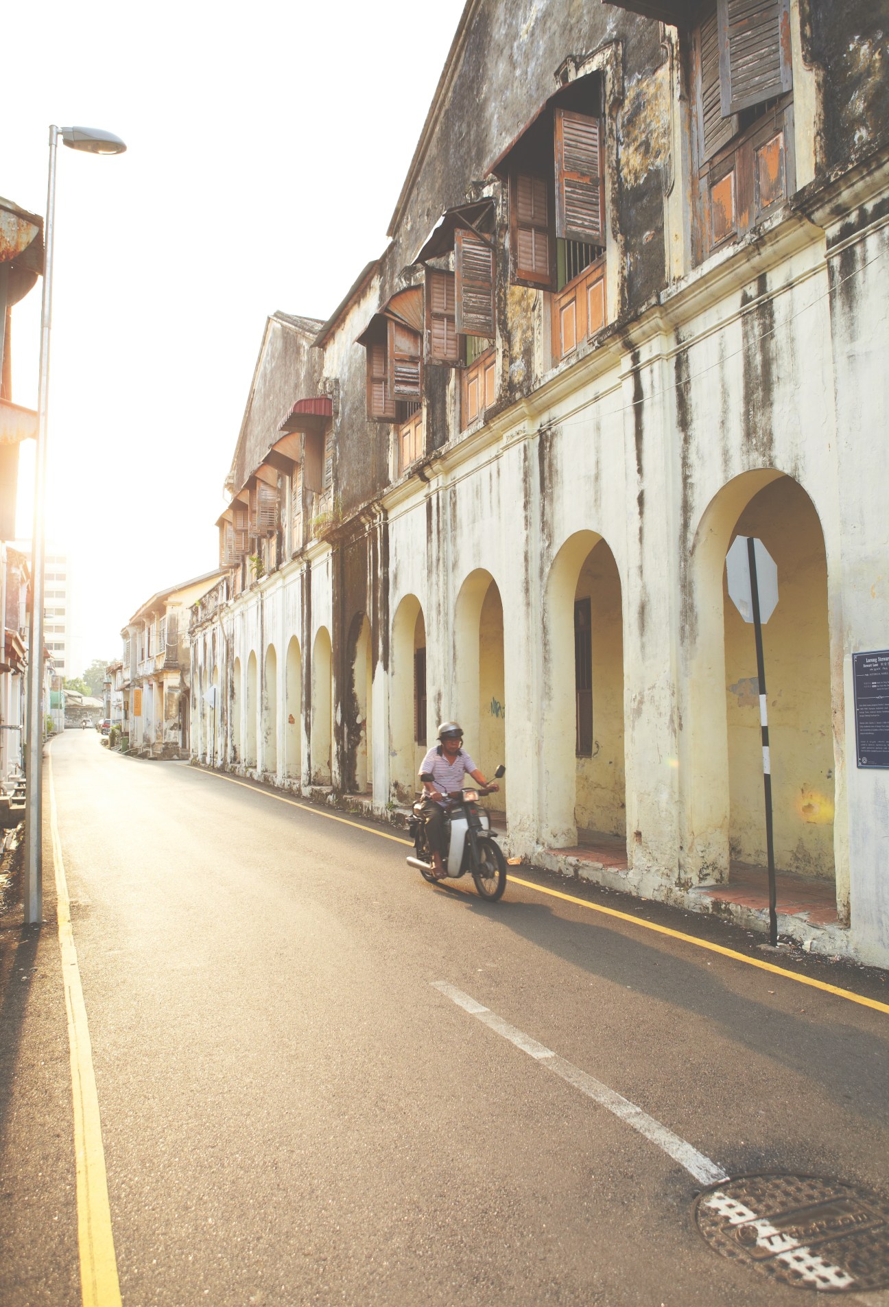 Person riding a motorcycle down a weathered colonial street with arched shop-house facades in George Town's Core Zone.