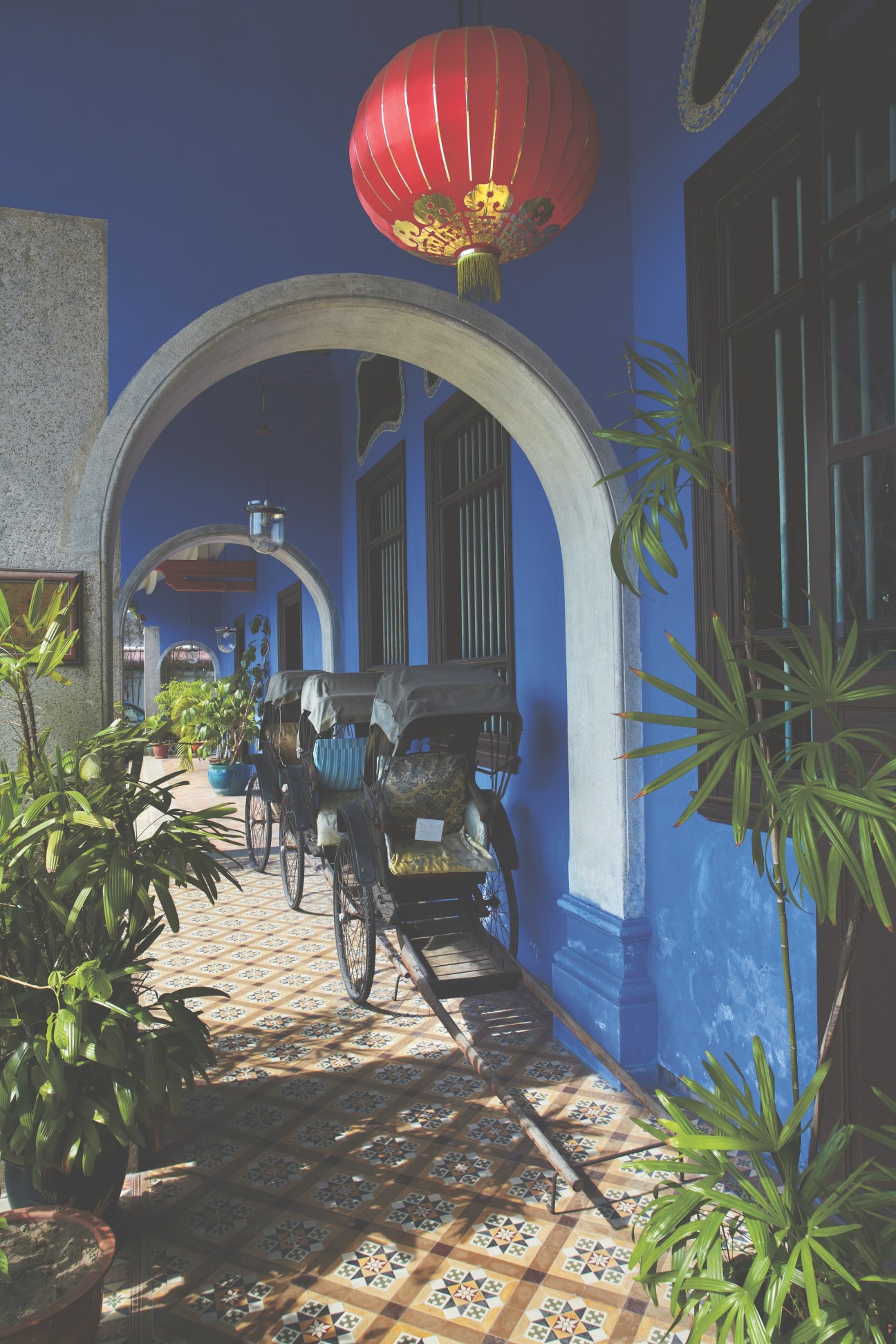 Blue-walled terrace with arched doorway, red lantern, decorative floor tiles and potted plants.