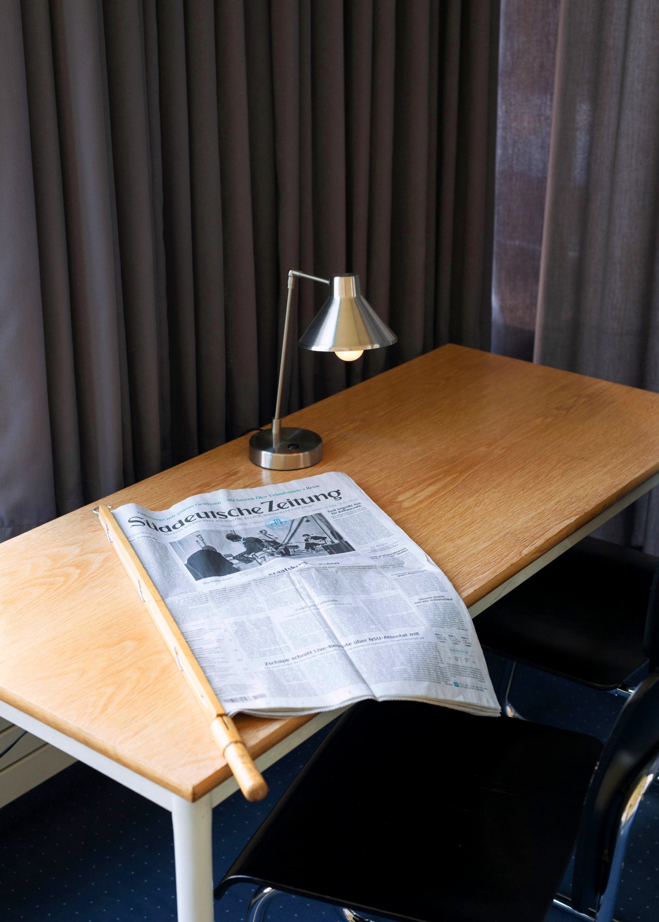 A wooden desk with a German newspaper and metal desk lamp against dark curtains.