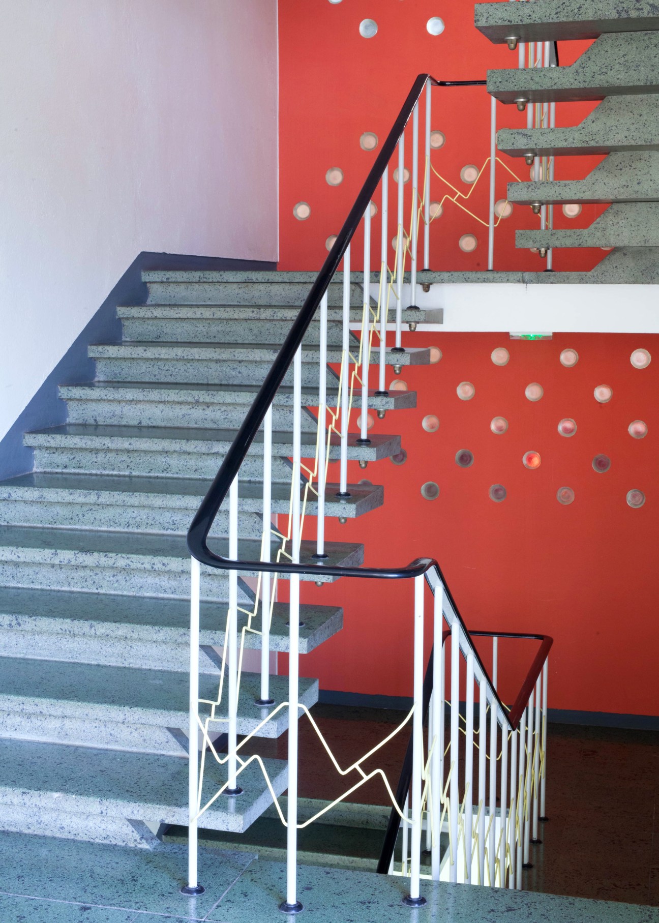 Concrete staircase with black handrail against red wall with circular perforations at Maison Heinrich Heine.