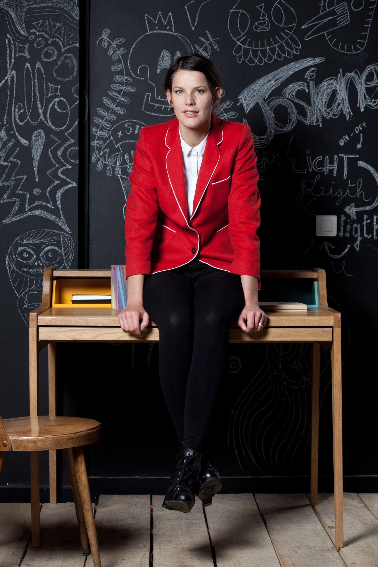 Woman in red blazer sitting on wooden desk with chalkboard background featuring hand-drawn designs.