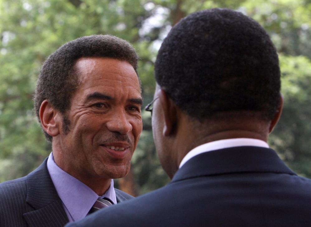 Two men in business suits conversing outdoors with green foliage in the background.