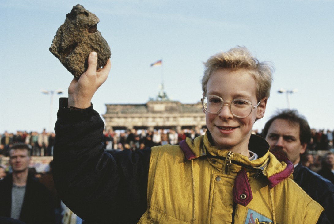 Young boy in yellow jacket and glasses holding a chunk of Berlin Wall stone at Brandenburg Gate.