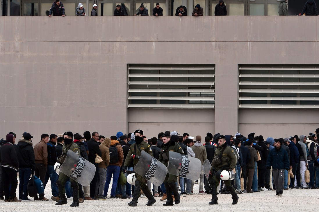 Riot police with shields standing between migrants and a concrete building wall in Athens.