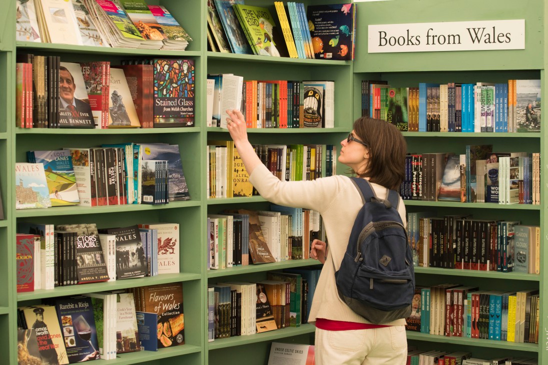 Woman browsing books on green shelves at a bookshop with a "Books from Wales" sign.