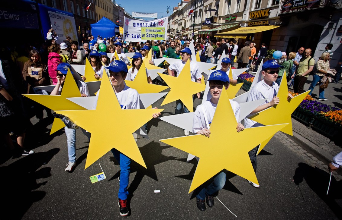 Polish children in white and blue clothing carrying large yellow EU stars during Europe Day parade in Warsaw.
