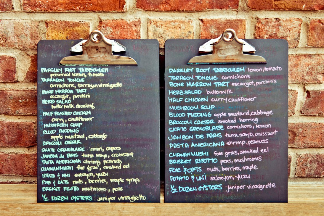 Two chalkboard menus with handwritten blue text mounted on a brick wall at M. Wells Dinette.