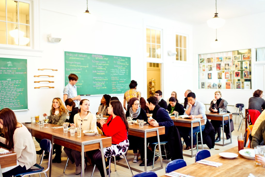 Diners seated at classroom-style wooden tables in M. Wells Dinette with green chalkboard menus on the walls.