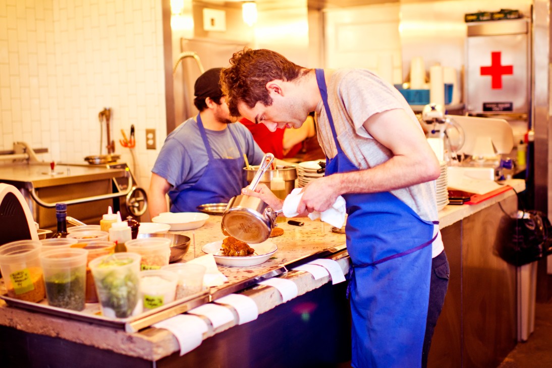Chefs working in a kitchen at M. Wells restaurant, plating food with various ingredients visible on the counter.