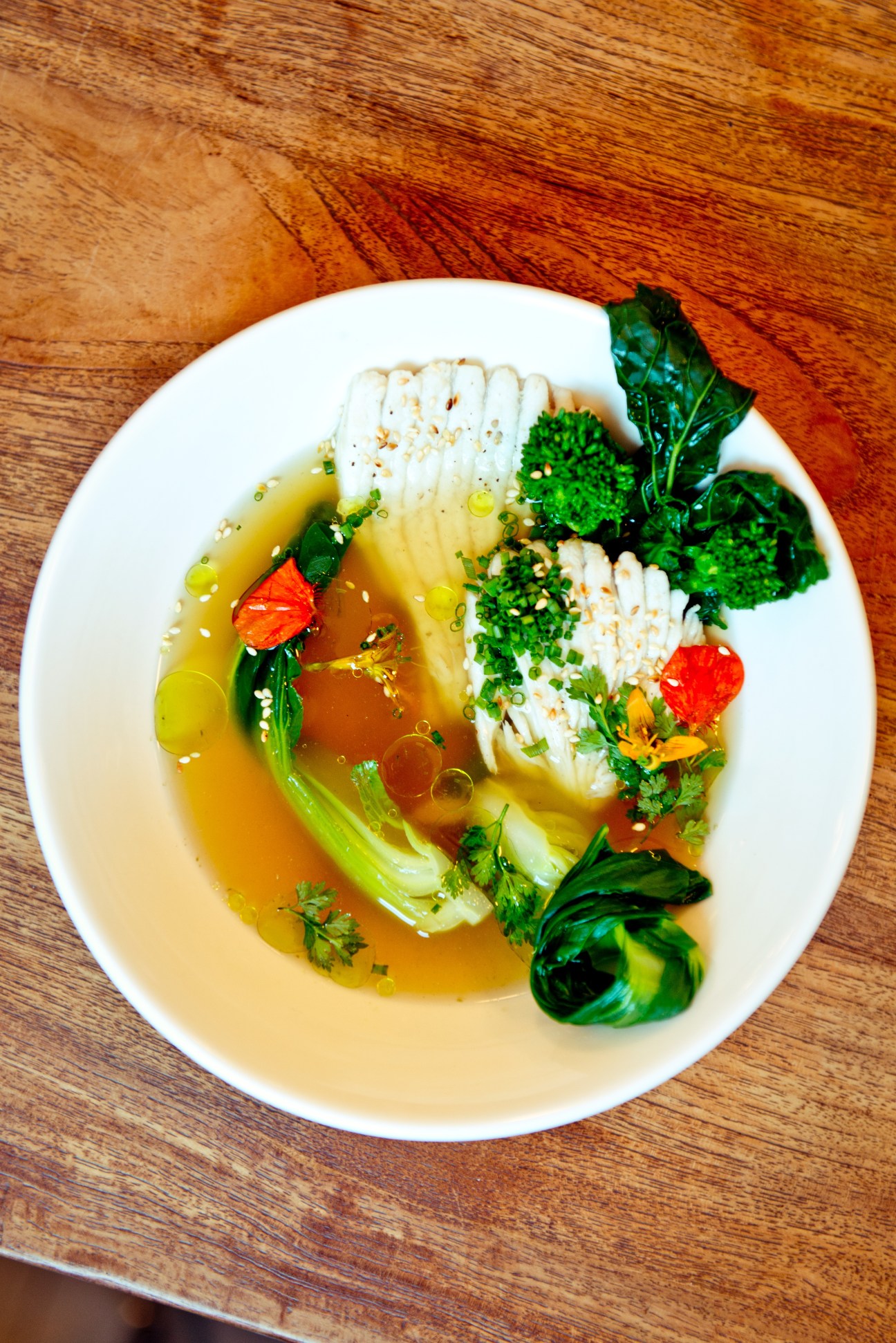 Skate poached in dashi broth with green vegetables and red pepper garnish on a white plate.