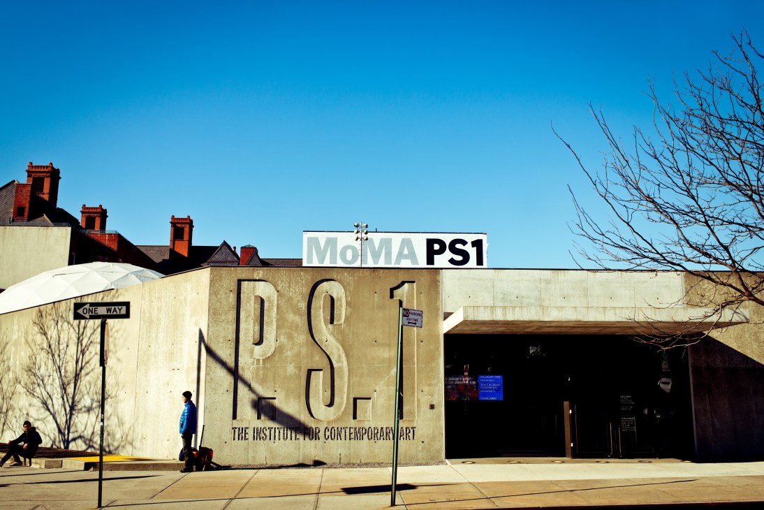 The award-winning entrance to MoMA PS1 with large lettering on a concrete facade under clear blue sky.
