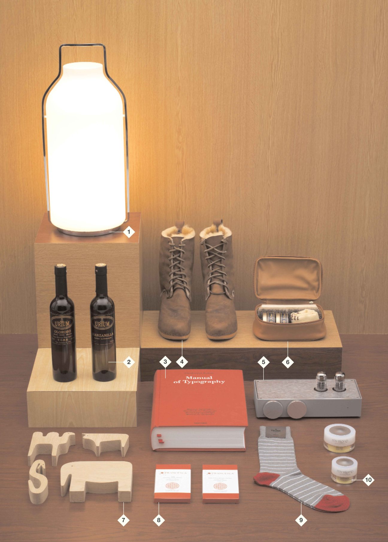 Styled gift guide arrangement featuring a glowing bottle lamp, wine bottles, boots, books, and accessories on wooden blocks.